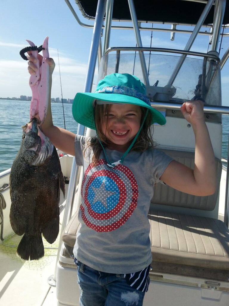 Young girl celebrating with a tripletail fish caught in Sarasota Bay, holding it up with pink grippers and pumping her fist in triumph